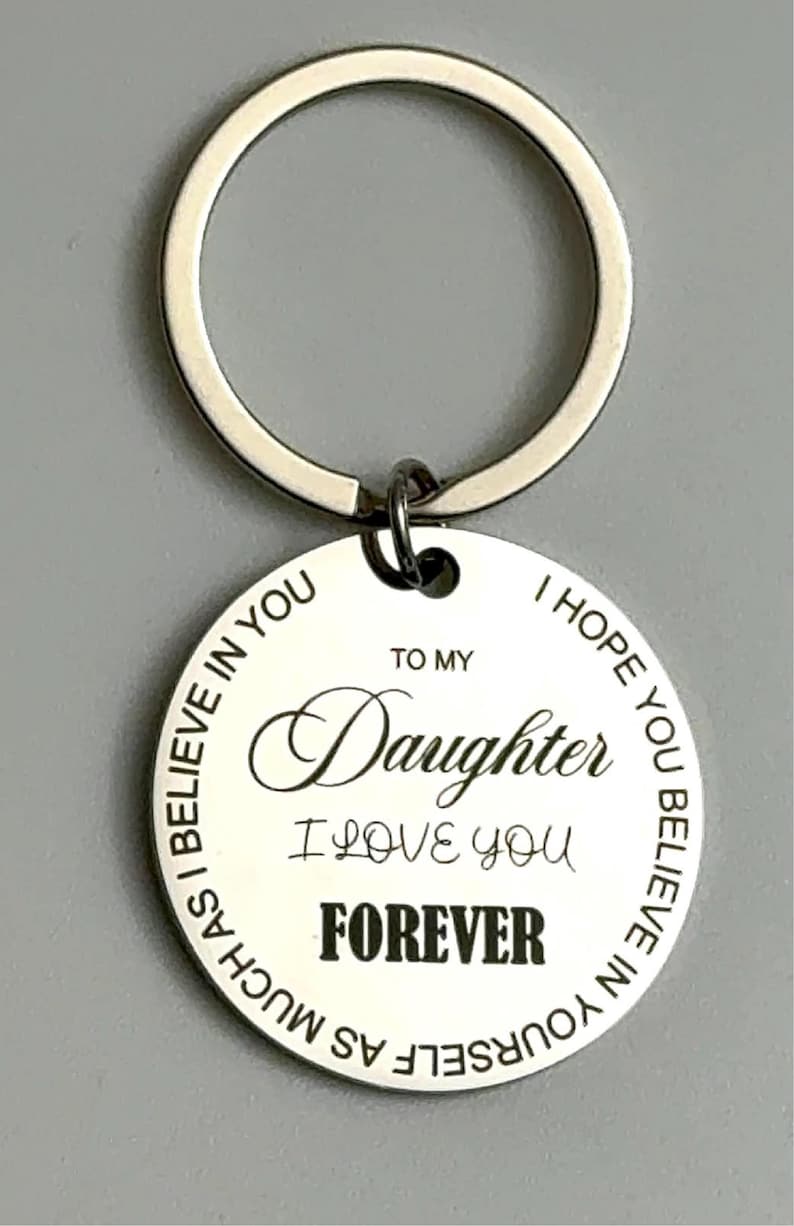 Close-up of a round silver keychain with a white engraved pendant. The pendant reads “To my Daughter I love you forever” in mixed script and bold fonts, with a circular border text that says, “I hope you believe in yourself as much as I believe in you.”