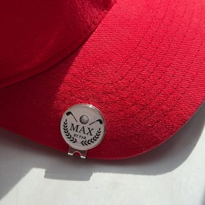 Puede incluir: Una gorra de b&eacute;isbol de color rojo brillante con un marcador de pelota de golf circular de metal plateado sujeto a la visera. El marcador muestra un gr&aacute;fico negro de una pelota de golf, dos palos cruzados y hojas de laurel con el texto MAX BY PAR.