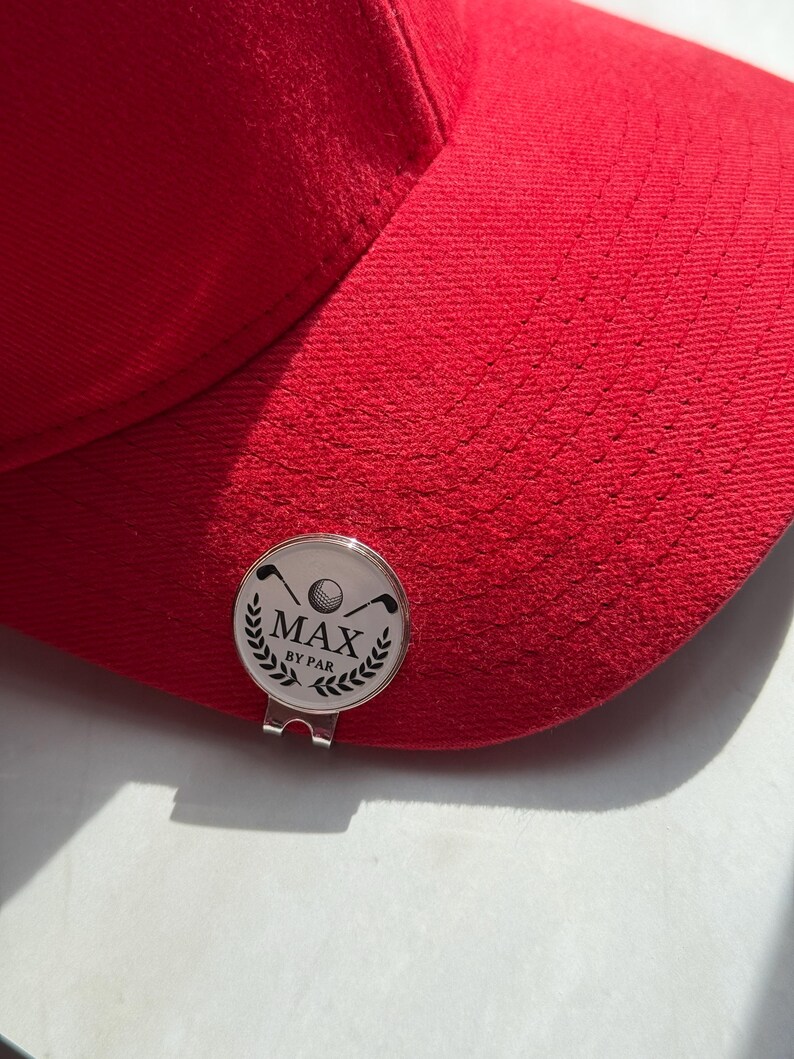 Puede incluir: Una gorra de b&eacute;isbol de color rojo brillante con un marcador de pelota de golf circular de metal plateado sujeto a la visera. El marcador muestra un gr&aacute;fico negro de una pelota de golf, dos palos cruzados y hojas de laurel con el texto MAX BY PAR.