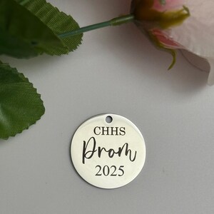 May include: A silver-colored, round charm with a hole at the top. The charm has the text "CHHS Prom 2025" in a black, elegant font. The background is a light gray color, with a pink flower and green leaves in the upper left corner.