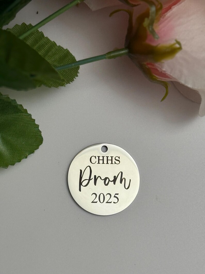 May include: A silver-colored, round charm with a hole at the top. The charm has the text "CHHS Prom 2025" in a black, elegant font. The background is a light gray color, with a pink flower and green leaves in the upper left corner.