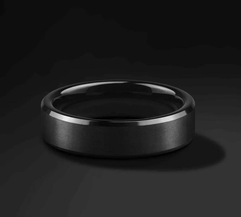 A black ring with a matte finish and a polished, beveled edge. The ring is set against a dark background, highlighting its sleek, modern design. This piece of jewelry is suitable for everyday wear.