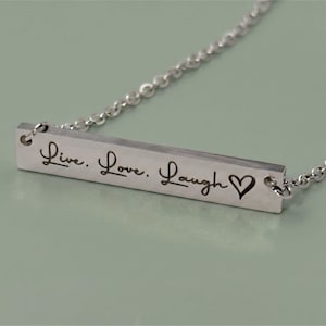 A silver-colored necklace with a rectangular pendant. The pendant is engraved with the words "Live. Love. Laugh" and a heart symbol. The necklace chain is delicate and made of small, linked pieces.