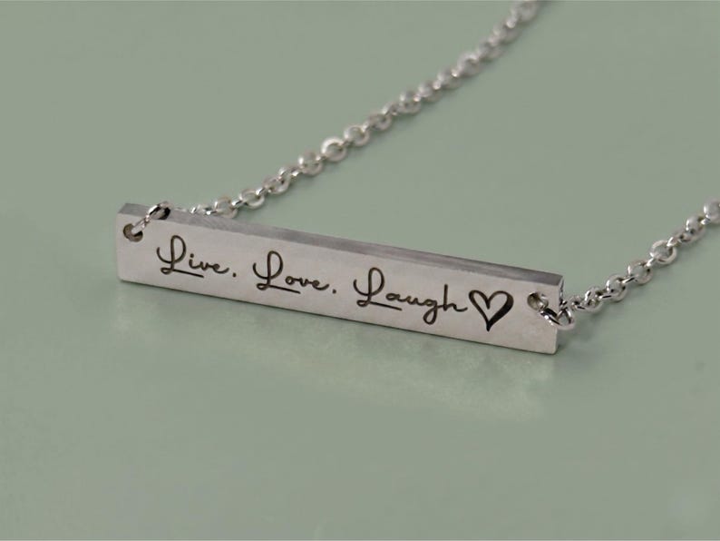 A silver-colored necklace with a rectangular pendant. The pendant is engraved with the words "Live. Love. Laugh" and a heart symbol. The necklace chain is delicate and made of small, linked pieces.