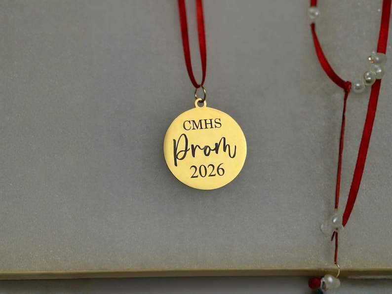 K&ouml;nnte beinhalten: Ein kreisf&ouml;rmiger goldfarbener Metallanh&auml;nger, der an einem d&uuml;nnen roten Band h&auml;ngt. Der Anh&auml;nger ist mit dem Text CMHS, dem Wort Prom in einer Schreibschrift und dem Jahr 2026 graviert. Ein zweites rotes Band mit kleinen wei&szlig;en Perlen ist rechts.