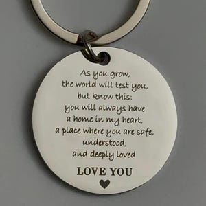 Close-up of a round silver keychain with a white engraved pendant featuring a heartfelt message about love, safety, and always having a home in the giver’s heart, ending with “LOVE YOU” and a small heart symbol.
