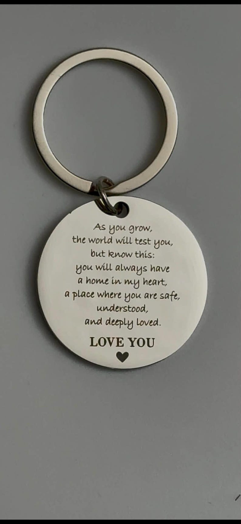 Close-up of a round silver keychain with a white engraved pendant featuring a heartfelt message about love, safety, and always having a home in the giver’s heart, ending with “LOVE YOU” and a small heart symbol.