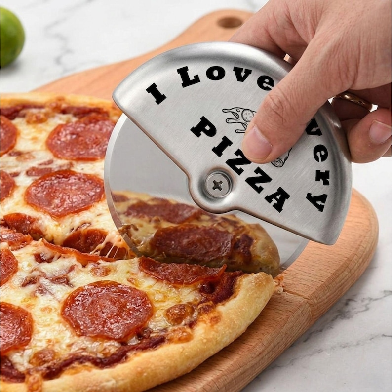 Close-up of a hand using a stainless steel pizza cutter engraved with “I Love Pizza” to slice a pepperoni pizza on a wooden cutting board, with melted cheese and crispy crust visible.