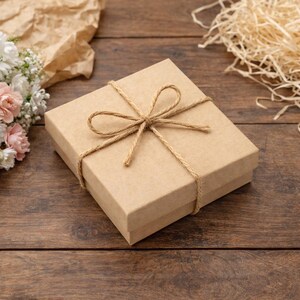 K&ouml;nnte beinhalten: Eine quadratische, hellbraune Geschenkbox, die mit Bindfaden gebunden ist und oben eine Schleife hat. Die Box liegt auf einer Holzoberfl&auml;che, mit einem Strau&szlig; wei&szlig;er und rosa Blumen und dekorativem Stroh in der N&auml;he. Das Gesamtthema ist das Schenken.