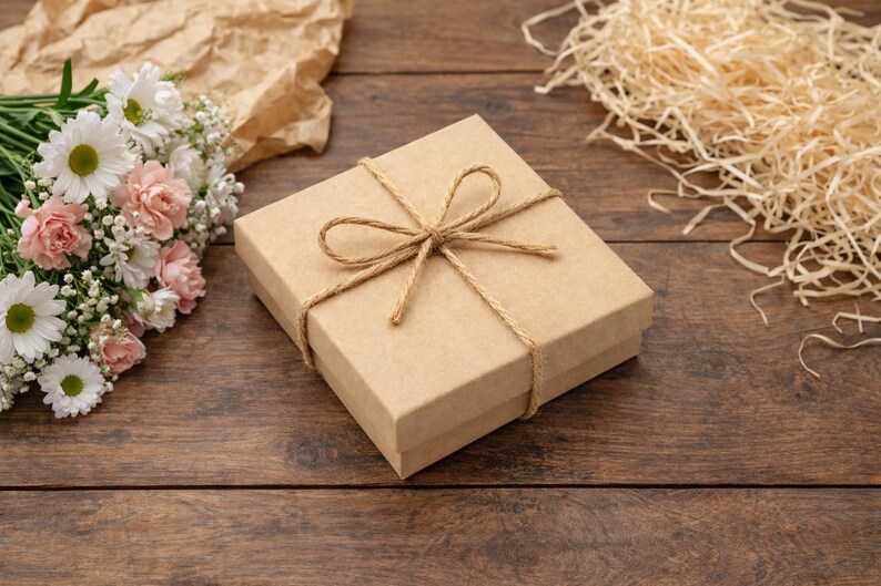 K&ouml;nnte beinhalten: Eine quadratische, hellbraune Geschenkbox, die mit Bindfaden gebunden ist und oben eine Schleife hat. Die Box liegt auf einer Holzoberfl&auml;che, mit einem Strau&szlig; wei&szlig;er und rosa Blumen und dekorativem Stroh in der N&auml;he. Das Gesamtthema ist das Schenken.
