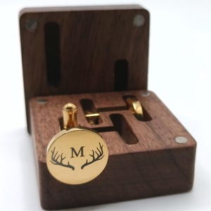 Puede incluir: Un gemelo dorado con la letra "M" y un dise&ntilde;o de cuernos de ciervo, presentado en una caja de madera marr&oacute;n oscuro abierta. La caja tiene ranuras para gemelos y un anillo. Los gemelos son redondos y brillantes.
