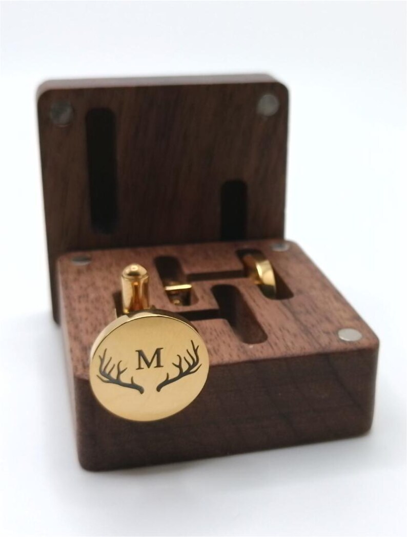 Puede incluir: Un gemelo dorado con la letra "M" y un dise&ntilde;o de cuernos de ciervo, presentado en una caja de madera marr&oacute;n oscuro abierta. La caja tiene ranuras para gemelos y un anillo. Los gemelos son redondos y brillantes.