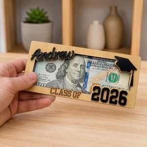 A hand holding a personalized laser-engraved wooden graduation money holder reading "Andrew – Class of 2026," with a $100 bill visible inside the slot, featuring a black 3D graduation cap accent on the corner, displayed against a warm wood table and blurred shelf background.