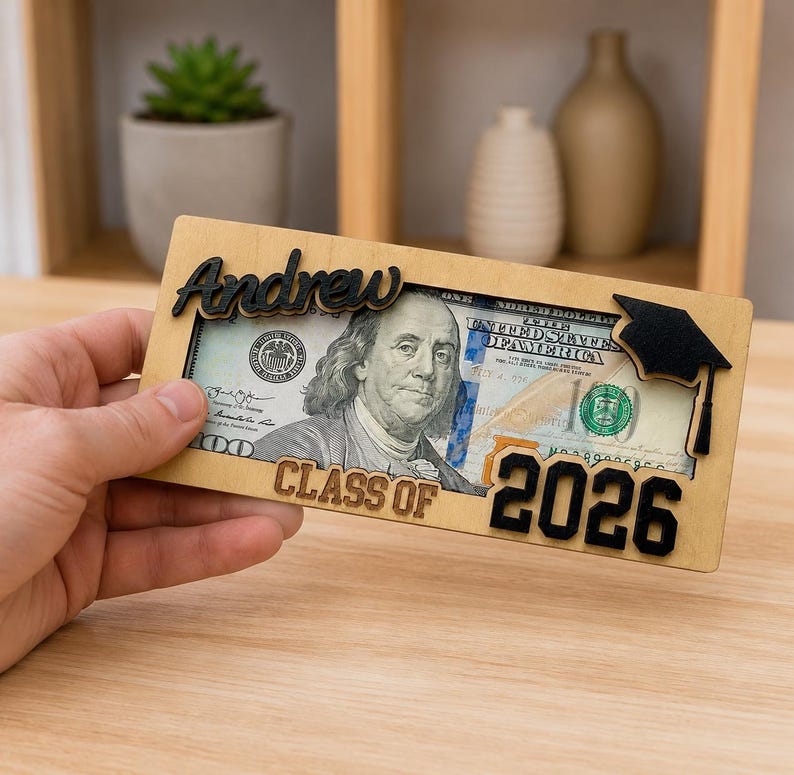 A hand holding a personalized laser-engraved wooden graduation money holder reading "Andrew – Class of 2026," with a $100 bill visible inside the slot, featuring a black 3D graduation cap accent on the corner, displayed against a warm wood table and blurred shelf background.
