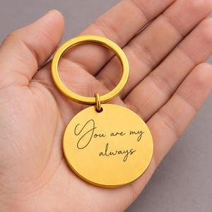 K&ouml;nnte beinhalten: Ein goldfarbener, kreisf&ouml;rmiger Metallschl&uuml;sselanh&auml;nger mit einer glatten, matten Oberfl&auml;che. Die Vorderseite des runden Anh&auml;ngers ist mit dem schwarzen kursiven Text "You are my always" graviert und an einem passenden goldenen Ring befestigt.