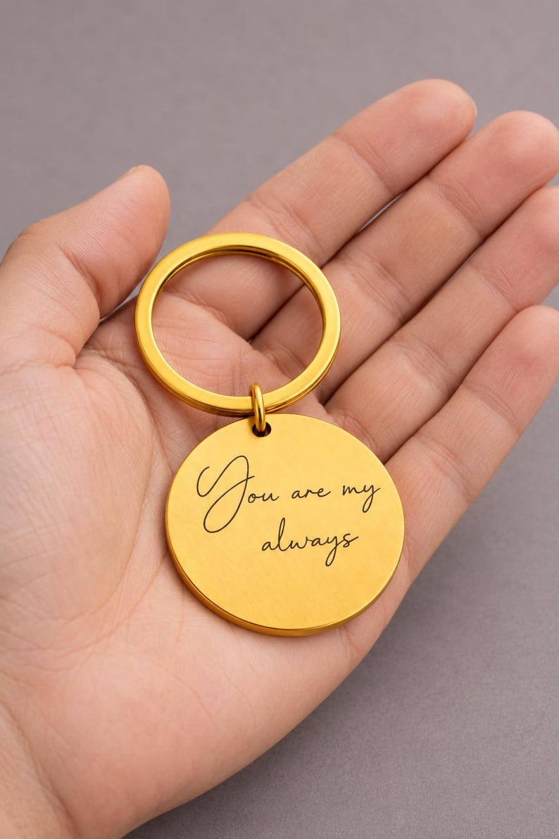 K&ouml;nnte beinhalten: Ein goldfarbener, kreisf&ouml;rmiger Metallschl&uuml;sselanh&auml;nger mit einer glatten, matten Oberfl&auml;che. Die Vorderseite des runden Anh&auml;ngers ist mit dem schwarzen kursiven Text "You are my always" graviert und an einem passenden goldenen Ring befestigt.