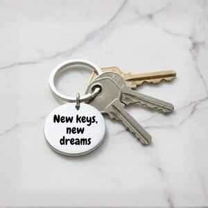 K&ouml;nnte beinhalten: Ein silberner Schl&uuml;sselanh&auml;nger mit einem runden Anh&auml;nger, auf dem in schwarzer Schrift "New keys, new dreams" steht. Der Schl&uuml;sselanh&auml;nger h&auml;lt drei Schl&uuml;ssel, zwei silberne und einen goldenen, vor einem Marmorhintergrund.