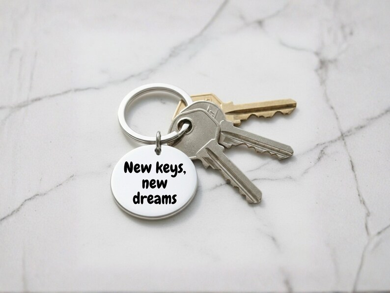 K&ouml;nnte beinhalten: Ein silberner Schl&uuml;sselanh&auml;nger mit einem runden Anh&auml;nger, auf dem in schwarzer Schrift "New keys, new dreams" steht. Der Schl&uuml;sselanh&auml;nger h&auml;lt drei Schl&uuml;ssel, zwei silberne und einen goldenen, vor einem Marmorhintergrund.