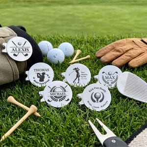 A collection of six personalized stainless steel golf ball markers with magnetic hat clips displayed on green grass. The custom engraved markers showcase various designs including names like Alexis, Thomas, Michael, Max, and custom text like "Best Dad Ever," "Best Mommy by Par," and "Kick Putt Daddy" alongside golf balls, tees, a glove, and a divot tool.