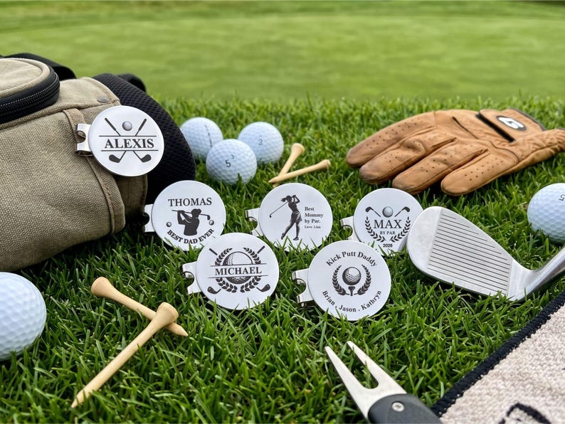 A collection of six personalized stainless steel golf ball markers with magnetic hat clips displayed on green grass. The custom engraved markers showcase various designs including names like Alexis, Thomas, Michael, Max, and custom text like "Best Dad Ever," "Best Mommy by Par," and "Kick Putt Daddy" alongside golf balls, tees, a glove, and a divot tool.