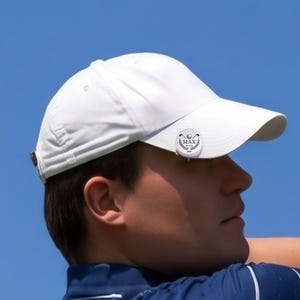 Close-up profile view of a male golfer wearing a white hat with a personalized magnetic golf ball marker clipped to the brim. The custom silver marker features an engraved laurel wreath logo, golf clubs, and the text "MAX BY PAR 2026" against a clear blue sky.