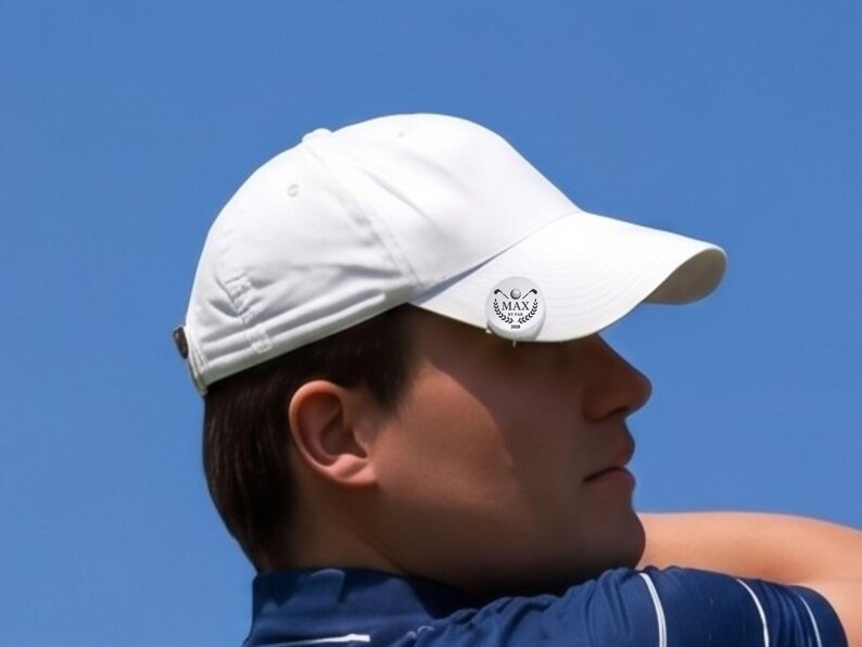 Close-up profile view of a male golfer wearing a white hat with a personalized magnetic golf ball marker clipped to the brim. The custom silver marker features an engraved laurel wreath logo, golf clubs, and the text "MAX BY PAR 2026" against a clear blue sky.