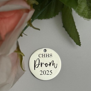 May include: A silver-colored, round charm with a hole at the top. The charm has the text "CHHS Prom 2025" in a black, elegant font. The background is a light gray color, with a pink flower and green leaves in the upper left corner.
