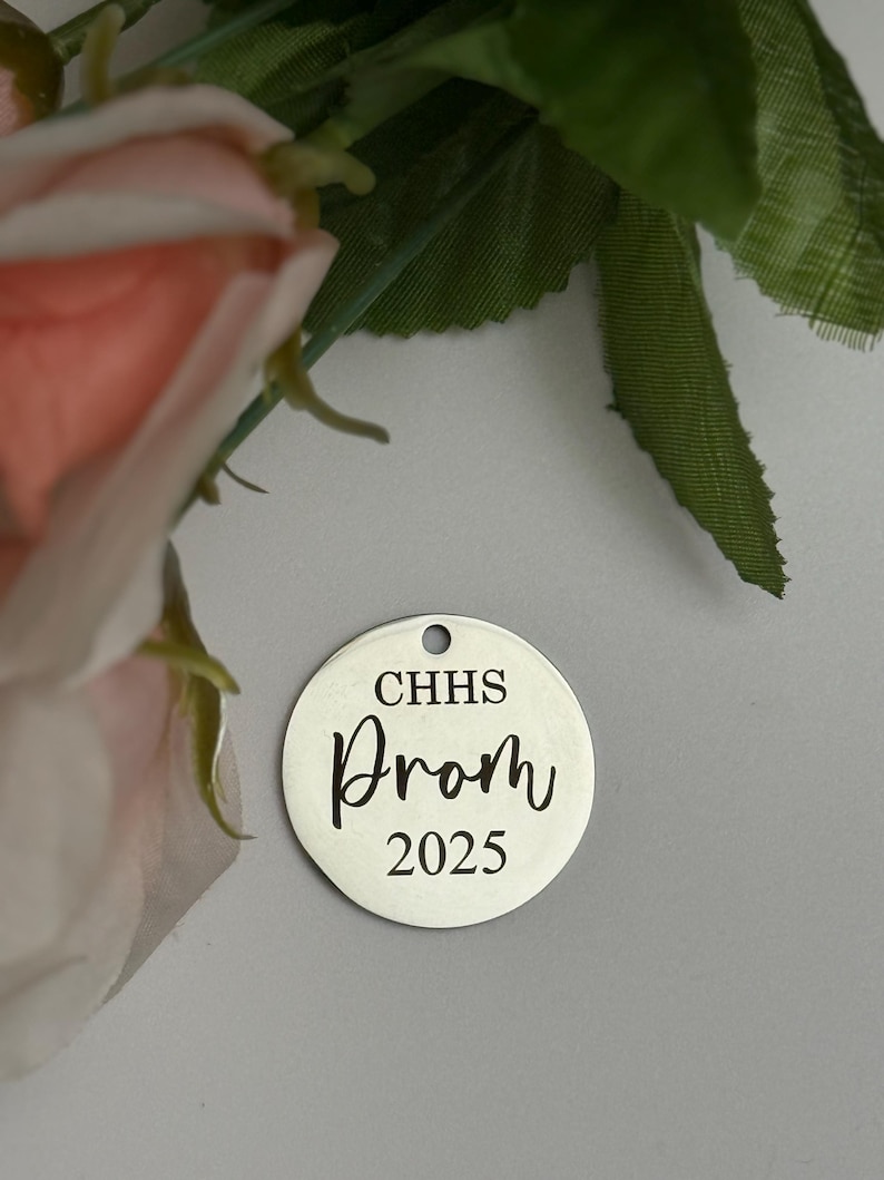 May include: A silver-colored, round charm with a hole at the top. The charm has the text "CHHS Prom 2025" in a black, elegant font. The background is a light gray color, with a pink flower and green leaves in the upper left corner.
