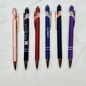 A set of six personalized metal ballpoint stylus pens displayed on a light marble surface. The pens come in assorted colors—rose gold, black, red, navy blue, matte black, and purple—each engraved with custom names or logos. Each pen features metallic accents, a stylus tip, and a sleek, modern design, perfect for engraving or gifting.