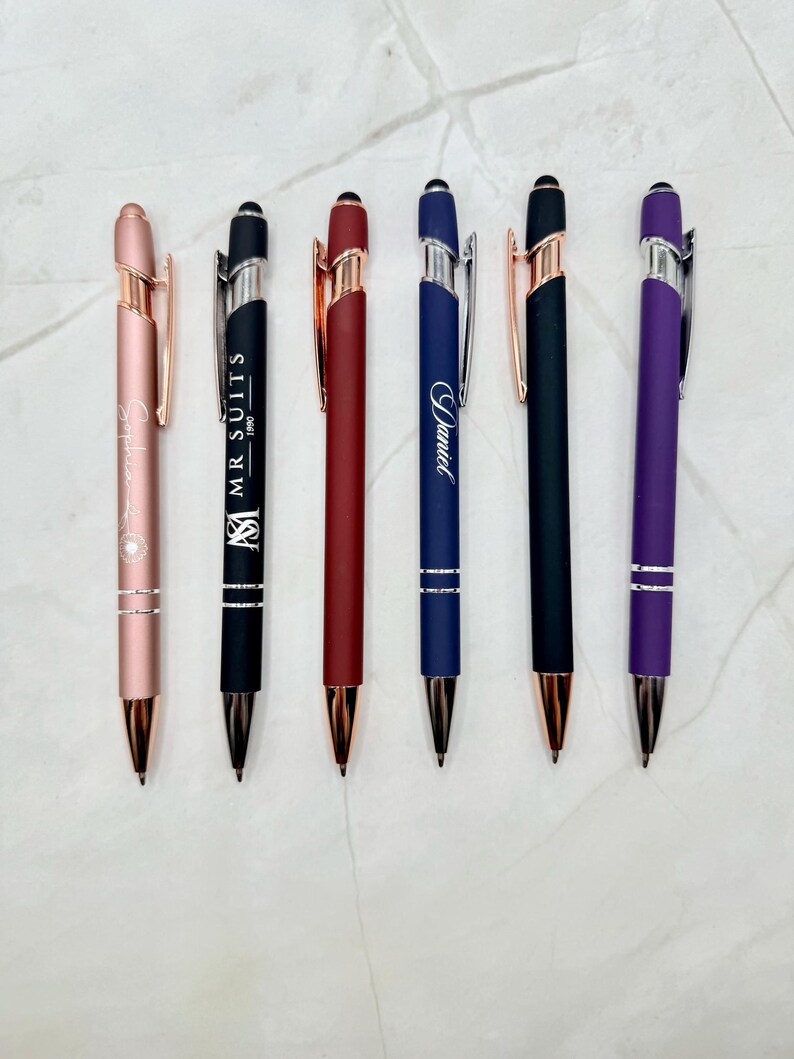 A set of six personalized metal ballpoint stylus pens displayed on a light marble surface. The pens come in assorted colors—rose gold, black, red, navy blue, matte black, and purple—each engraved with custom names or logos. Each pen features metallic accents, a stylus tip, and a sleek, modern design, perfect for engraving or gifting.