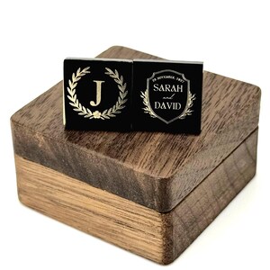 Two black square cufflinks with gold-colored designs rest on a wooden box. One cufflink features a gold laurel wreath around the letter "J". The other cufflink has the names "Sarah and David" and the date "28 November, 2021".