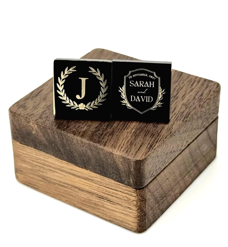 Two black square cufflinks with gold-colored designs rest on a wooden box. One cufflink features a gold laurel wreath around the letter "J". The other cufflink has the names "Sarah and David" and the date "28 November, 2021".