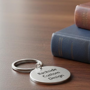 Round metal keychain with a brushed silver finish lying on a wooden table, engraved with the words “Backside Custom Design,” placed beside two stacked books.
