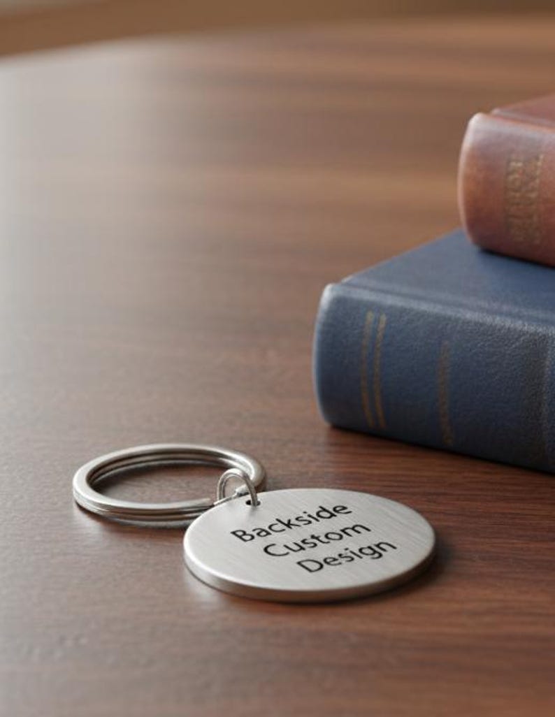 Round metal keychain with a brushed silver finish lying on a wooden table, engraved with the words “Backside Custom Design,” placed beside two stacked books.