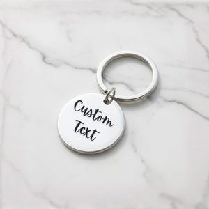Puede incluir: Un llavero plateado con un colgante redondo. El colgante tiene las palabras "Custom Text" en una fuente cursiva negra. El llavero est&aacute; sobre una superficie de m&aacute;rmol blanco y gris.