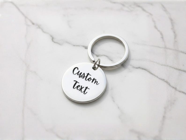 Puede incluir: Un llavero plateado con un colgante redondo. El colgante tiene las palabras "Custom Text" en una fuente cursiva negra. El llavero est&aacute; sobre una superficie de m&aacute;rmol blanco y gris.