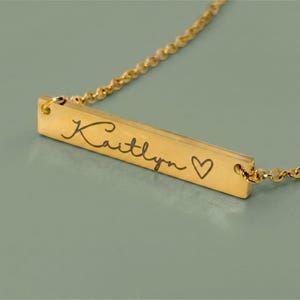 A gold-colored necklace with a rectangular pendant. The pendant is engraved with the name "Kaitlyn" in cursive script, followed by a small heart symbol. The necklace chain is delicate and also gold-colored.
