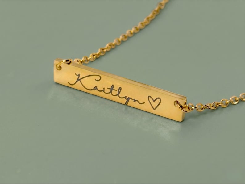 A gold-colored necklace with a rectangular pendant. The pendant is engraved with the name "Kaitlyn" in cursive script, followed by a small heart symbol. The necklace chain is delicate and also gold-colored.