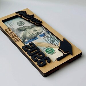 Low-angle side view of a personalized laser-engraved birch wood graduation money holder reading "Andrew – Class of 2026," revealing the multi-layer wood construction with a $100 bill tucked inside the slot, raised black script name on top, bold black laser-cut "2026" numbers, and a laser-cut graduation cap with tassel on the right, photographed against a clean white background.