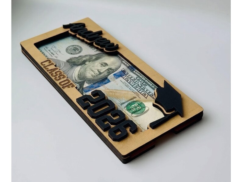 Low-angle side view of a personalized laser-engraved birch wood graduation money holder reading "Andrew – Class of 2026," revealing the multi-layer wood construction with a $100 bill tucked inside the slot, raised black script name on top, bold black laser-cut "2026" numbers, and a laser-cut graduation cap with tassel on the right, photographed against a clean white background.