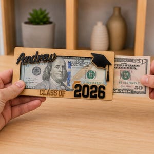 Two hands sliding a $50 bill into a personalized laser-engraved wooden graduation money holder reading "Andrew – Class of 2026," with a $100 bill already inside, featuring a black 3D graduation cap accent, displayed on a wood table with a cozy shelf background.