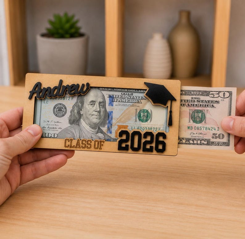 Two hands sliding a $50 bill into a personalized laser-engraved wooden graduation money holder reading "Andrew – Class of 2026," with a $100 bill already inside, featuring a black 3D graduation cap accent, displayed on a wood table with a cozy shelf background.