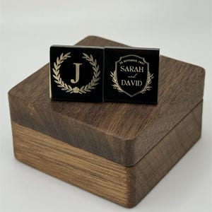 Puede incluir: Dos gemelos cuadrados negros con dise&ntilde;os dorados descansan sobre una caja de madera. Un gemelo presenta una corona de laurel dorada alrededor de la letra "J". El otro gemelo tiene los nombres "Sarah y David" y la fecha "28 de noviembre de 2021".