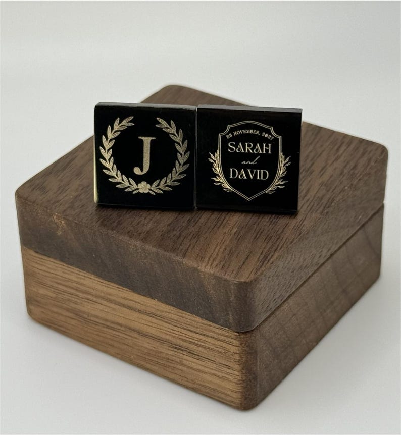 Puede incluir: Dos gemelos cuadrados negros con dise&ntilde;os dorados descansan sobre una caja de madera. Un gemelo presenta una corona de laurel dorada alrededor de la letra "J". El otro gemelo tiene los nombres "Sarah y David" y la fecha "28 de noviembre de 2021".