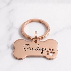 Personalized rose gold dog tag with engraved name Penelope in cursive. Custom bone-shaped brass pet ID tag with paw print cutout and gold keyring on a marble surface.