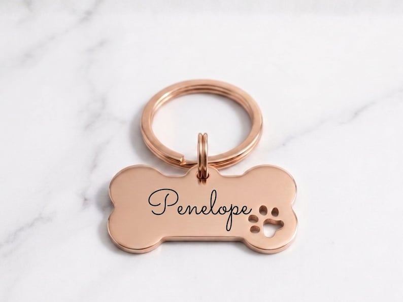 Personalized rose gold dog tag with engraved name Penelope in cursive. Custom bone-shaped brass pet ID tag with paw print cutout and gold keyring on a marble surface.
