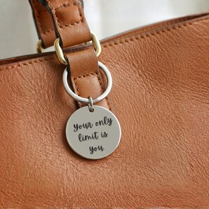 K&ouml;nnte beinhalten: Ein silberfarbener runder Schl&uuml;sselanh&auml;nger mit der Aufschrift "your only limit is you" h&auml;ngt an einer braunen Ledertasche. Die Tasche hat eine strukturierte Oberfl&auml;che und einen braunen Lederriemen mit goldfarbenen Metallbeschl&auml;gen.