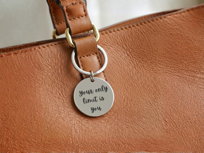 K&ouml;nnte beinhalten: Ein silberfarbener runder Schl&uuml;sselanh&auml;nger mit der Aufschrift "your only limit is you" h&auml;ngt an einer braunen Ledertasche. Die Tasche hat eine strukturierte Oberfl&auml;che und einen braunen Lederriemen mit goldfarbenen Metallbeschl&auml;gen.