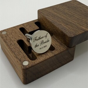 Puede incluir: Un gemelo plateado grabado con "Father of the Bride" y la fecha 10.11.2025, presentado en una caja de madera oscura. La caja tiene una tapa con bisagras y un interior personalizado para sujetar el gemelo de forma segura. El gemelo es redondo.