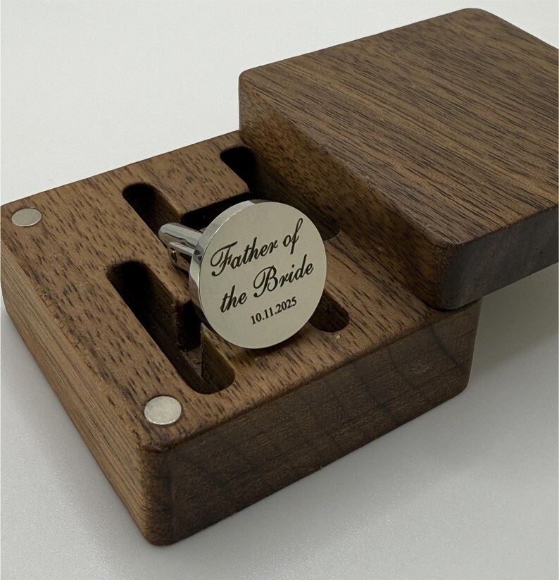 Puede incluir: Un gemelo plateado grabado con "Father of the Bride" y la fecha 10.11.2025, presentado en una caja de madera oscura. La caja tiene una tapa con bisagras y un interior personalizado para sujetar el gemelo de forma segura. El gemelo es redondo.